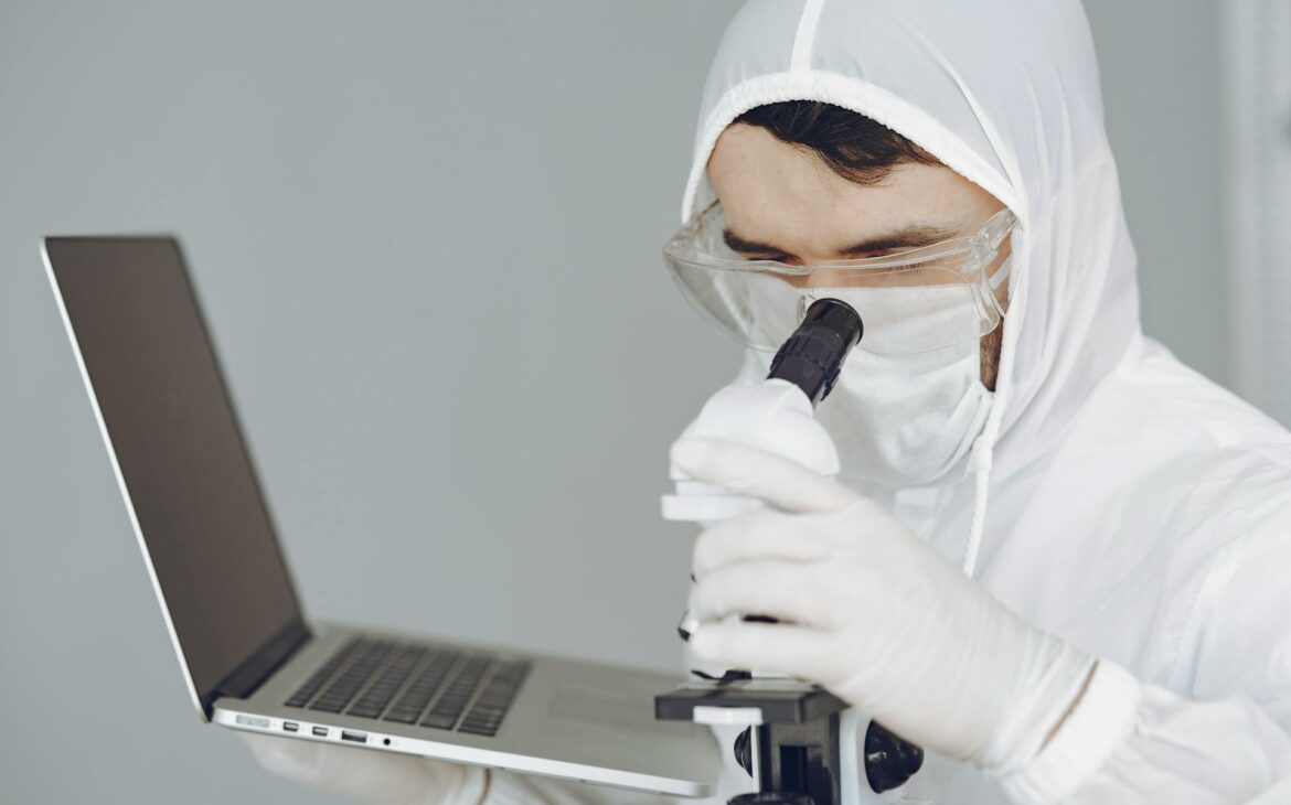  a scientist wearing personal protective equipment and holding a laptop looking through a microscope