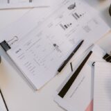Documents and charts spread on a desk with a pen, notebooks, and coffee cups