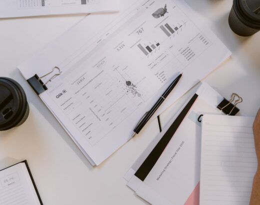 Documents and charts spread on a desk with a pen, notebooks, and coffee cups