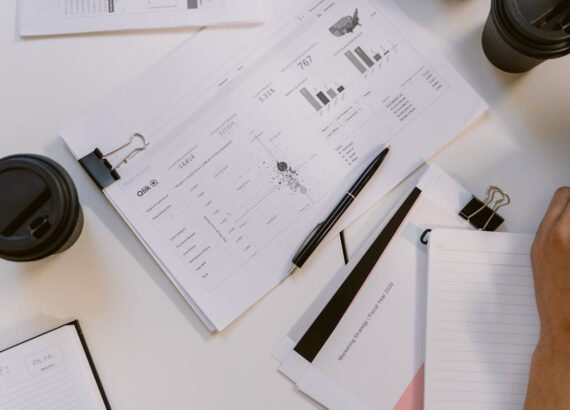Documents and charts spread on a desk with a pen, notebooks, and coffee cups